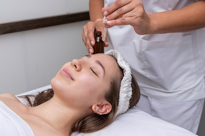 A young woman lays on a procedure table with a licensed ethsthetican performs a dermaplaning procedure